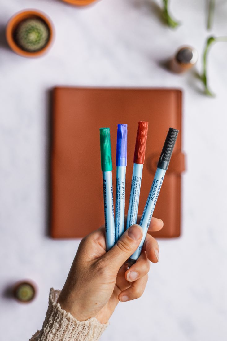 Hand holding erasable pens in green, blue, red and black with brown vegan leather cover notebook in the background
