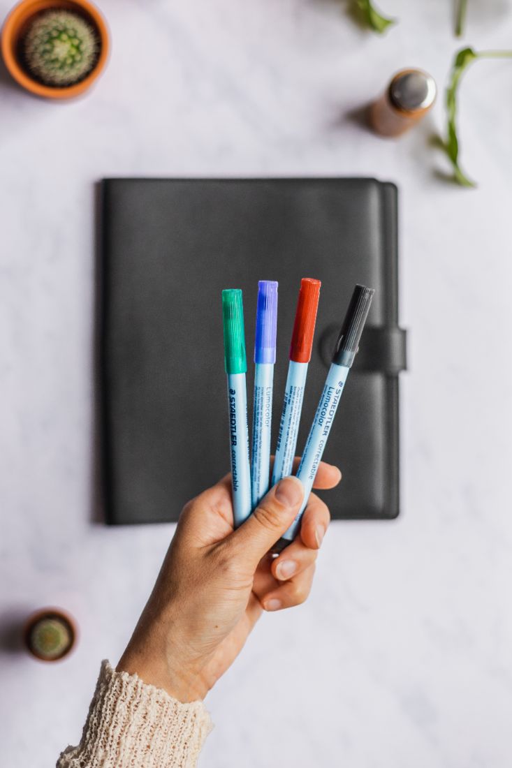 Hand holding erasable pens in green, blue, red and black with black vegan leather cover notebook in the background