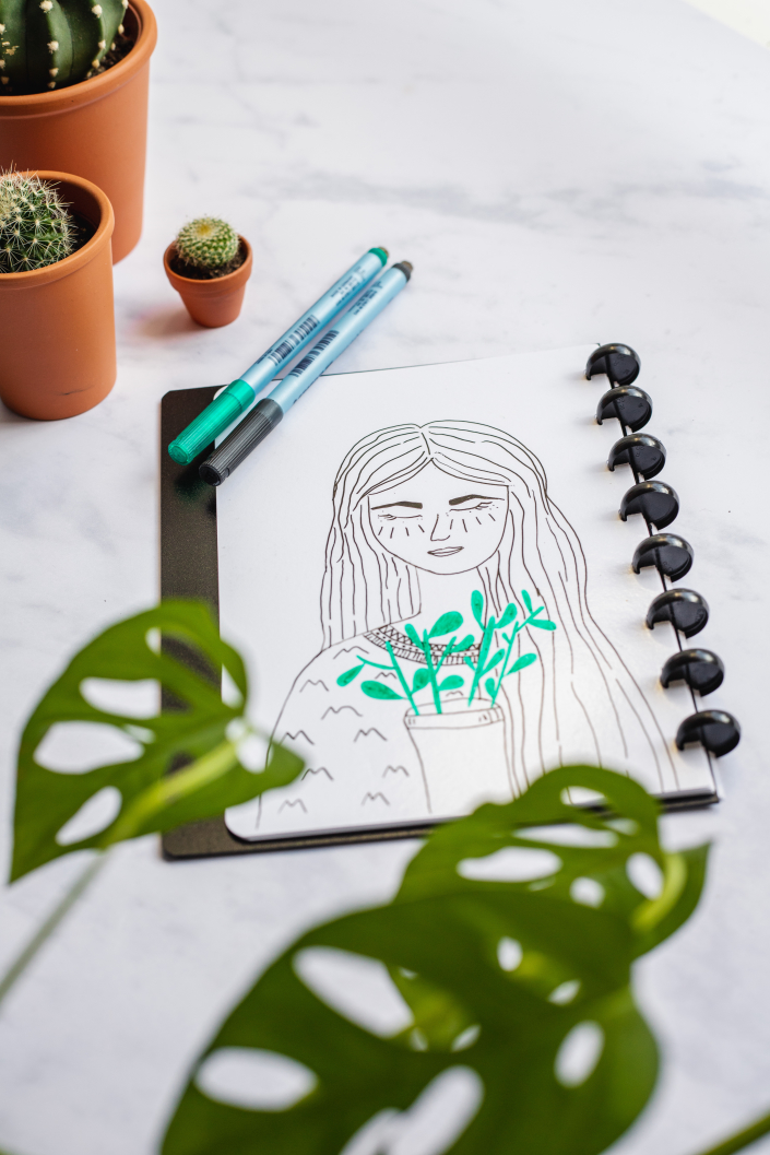 Drawing of woman and plant in notebook with plants surrounding on a marble background