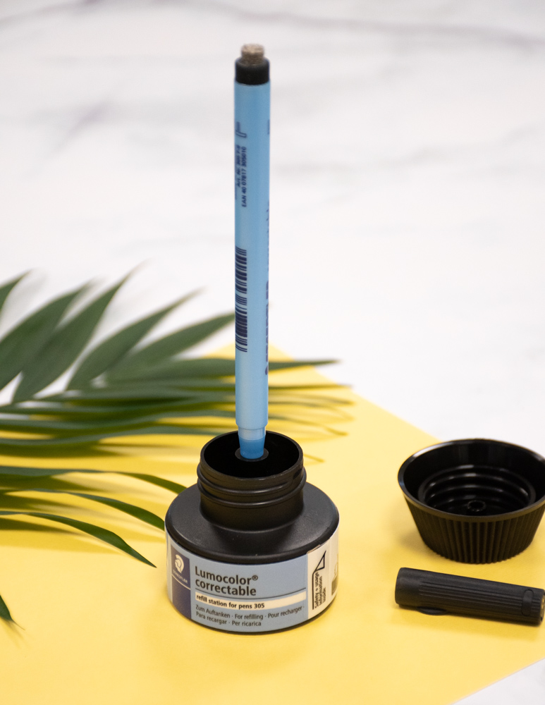 Erasable pen sticking upright in black ink refill station on yellow paper with plant