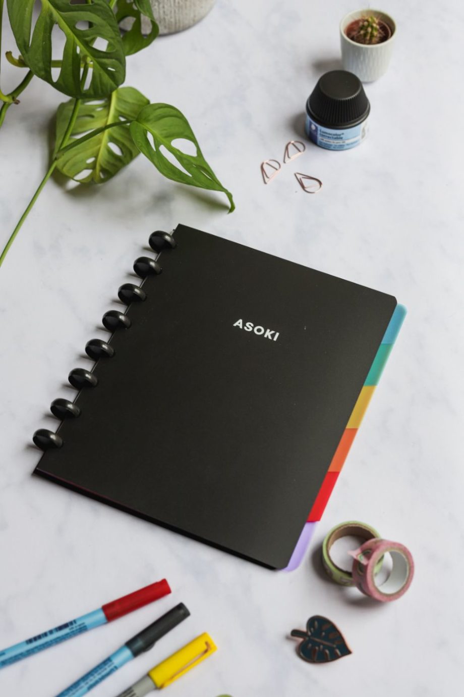 Black notebook with white letters and colourful dividers next to assorted items on marble background