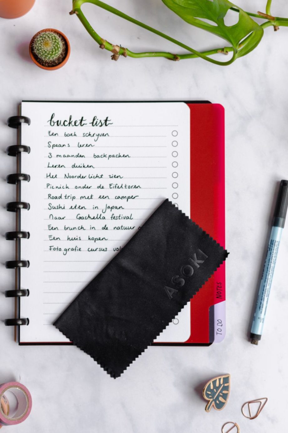 To-do list notebook with black ringbinding and Asoki black wipe on top with pen on assorted items on marble background