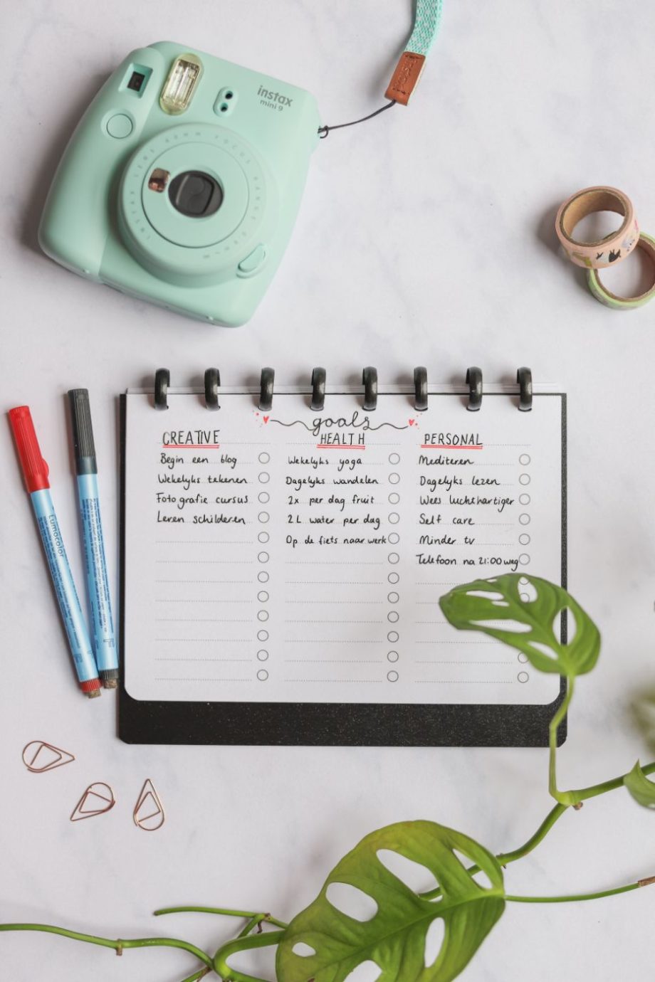 Horizontal erasable filled-in to-do list with black rings and black cover next to pens, camera and plant on marble background