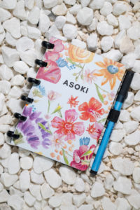 A6 reusable notebook with red, purple, yellow and blue flowers with green stems on a light blue background. On the right side of the A6 reusable notebook, a black elastic pen loop is visible with a pen in it. The pen is about as long as the notebook is.